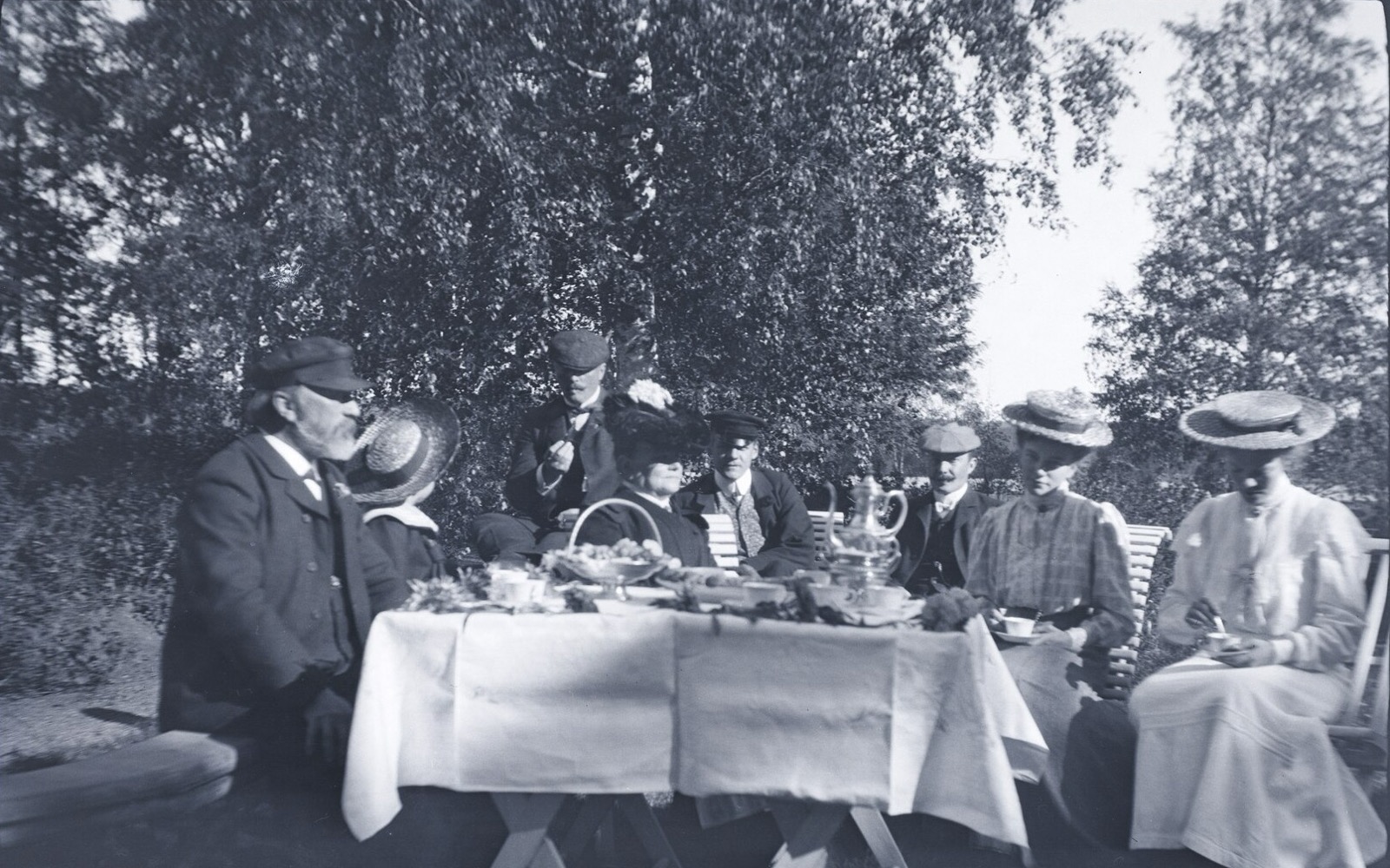 En grupp kvinnor och män i hattar dricker kaffe vid ett dukat bord i en trädgård